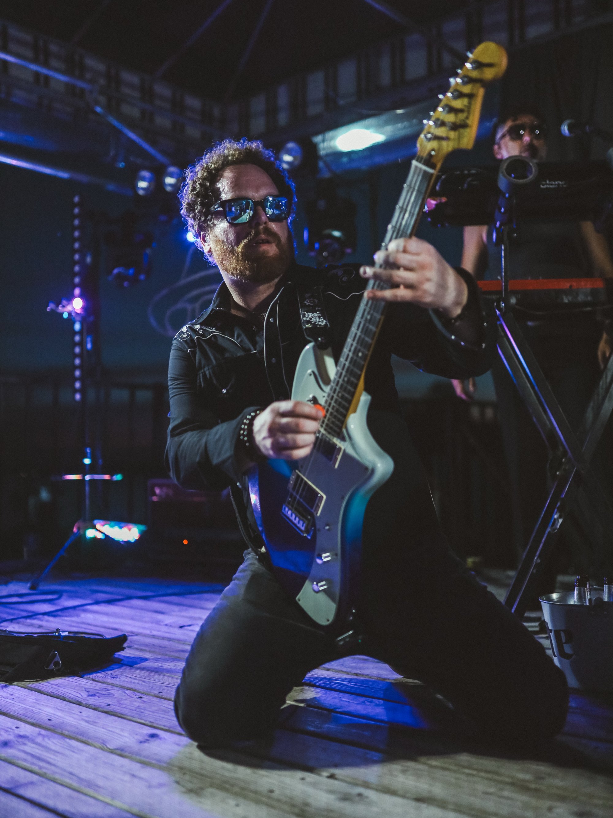 Dave Menzo on one knee on stage with a guitar, blue light