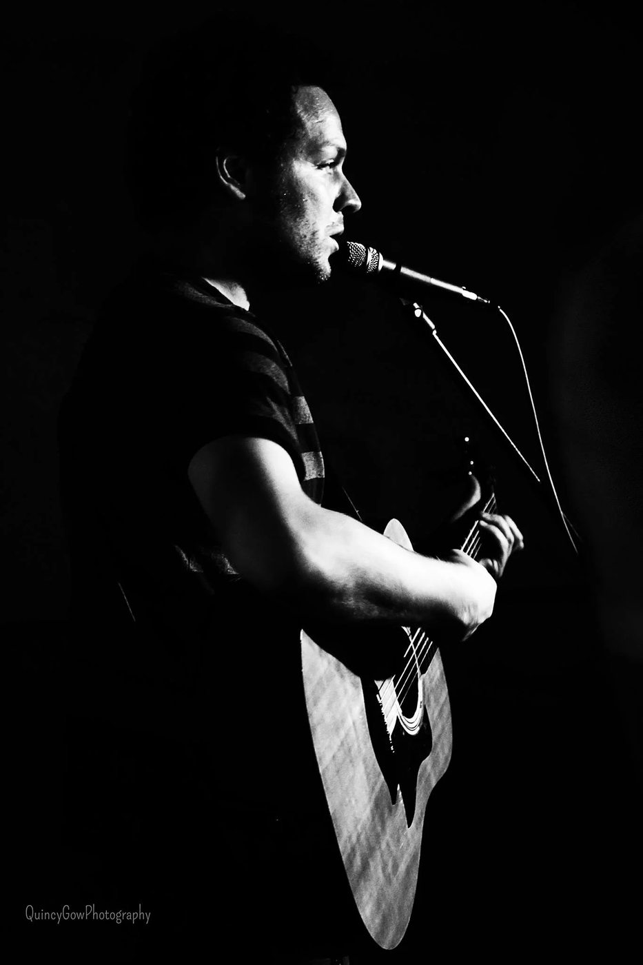 Dave Menzo singing into a microphone with an acoustic guitar, lit from the side on a dark stage