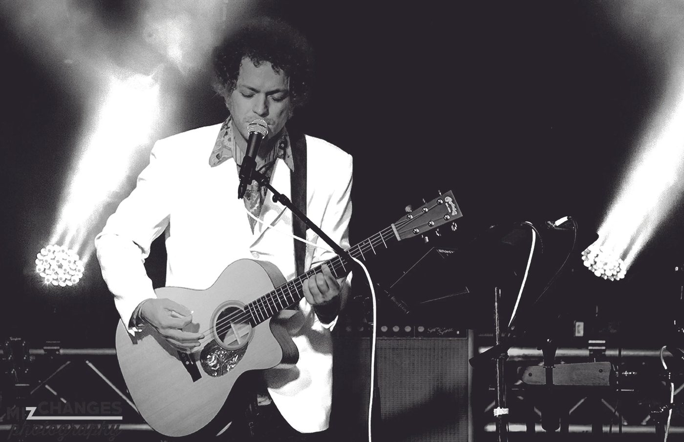 Dave Menzo playing acoustic guitar under white stage lights, black and white